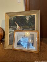 Two framed artworks leaning against a wall; one depicts a man fishing by a river with a larger wood frame, the other a smaller framed sailing ship with a city in background.