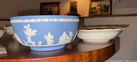 Blue Wedgwood jasperware bowl and white ceramic floral bowl side by side on a wooden surface with framed pictures in the background.