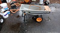 Full side view angle of the Worx WG050 wheelbarrow with visible orange handles and wheels, metal tray, and black frame. Some rust noted on tray and frame.