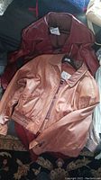 Two women's leather jackets lying flat on a dark surface, one reddish-burgundy, one light brown.
