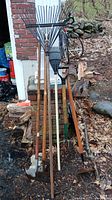 Photo of six yard tools including two edgers, hoe with broken handle, two grass rakes, and garden fork leaning against brick background outdoors