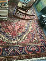 Top-down view of large rectangular area rug with red and navy blue floral and ornate designs beneath a rocking chair and stacked books.