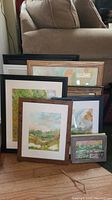 Photo showing eleven framed artworks leaning against a chair, varying in sizes and frame colors from black to wooden tones, depicting landscapes and abstract scenes.