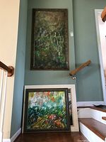 Three framed paintings displayed near a staircase, one large vertical above two smaller square paintings stacked on floor.