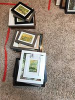 Top view of multiple stacked framed art pieces on carpet, showing a variety of frames and artwork sizes