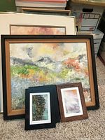 Grouped framed art pieces stacked against wall on carpet with various sizes and frames.