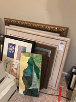 Photo showing a stacked assortment of framed artworks of varying sizes and frame styles, including ornate gold frame, plain white frame, and black frame, as well as a vertically oriented elongated canvas with abstract green and earth tones.