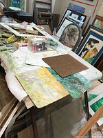 6 foot folding catering table loaded with painting materials and artwork, surface visibly stained.