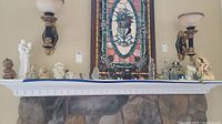 Mantel shelf with multiple figurines including porcelain couples, angels, and other figurines alongside two glass and metal wall sconces and a stained glass panel