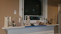 Full view of various figurines including birds, people, musicians, and a typewriter on a shelf beneath a television, showing quantity and variety.