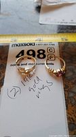 Two gold rings placed on paper with handwritten notes and measurement tape showing sizes and length, one is the Claddagh ring and the other has a purple gemstone.