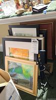 Stack of framed art pieces leaning against a wall, showing a variety of frame styles and sizes with a visible price tag on one frame.