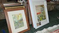 Photo shows multiple framed paintings stacked on carpet, showcasing two pieces with distinct frames and matting