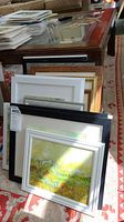Stack of 15 framed paintings leaning against a table, showing frame variety and size range