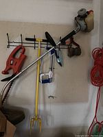 Overall view of several yard tools hanging on a wall including the edger, hedge trimmer, garden fork, hand aerator, and saw.