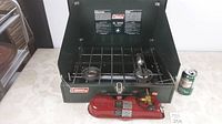 Green Coleman 2-burner camp stove with metal grate on top and red fuel tank in front. A soda can is beside it for scale.