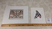 Two Haida art prints side by side, showing designs in black and red traditional Indigenous style, with a measuring ruler beneath the frames.