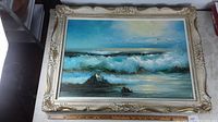 Full view of the framed oil painting showing the ornate cream decorative frame with sculpted detailing and the seascape art inside.