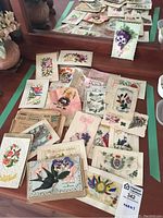 Wide view of collection of antique embroidered greeting cards and handkerchiefs arranged on a surface with background reflection from mirror, showing various floral motifs and delicate paper lace work.