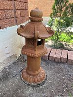 Full view of rust-colored Asian garden statue with curved roof and wide base with petal shapes in garden dirt near house foundation.