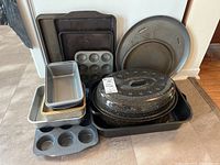 Assorted bakeware including roaster with lid, baking sheets, pizza pans, and cupcake trays stacked against wall.