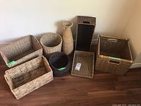 Full set of 7 woven baskets and trays arranged on a wooden floor against a beige wall showing their shapes and sizes.