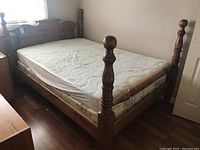 Four-poster wooden bed frame with mattress and box spring on a hardwood floor near a window. Shows scratches on the wood frame and fabric mattress and box spring.