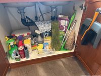 Cleaning supplies and tools stored under kitchen sink, showing various spray bottles, dusters, brushes, sponges and cleaning chemicals