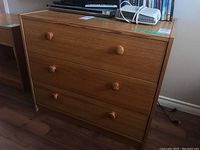 Front-facing view of laminate dresser showing three drawers with round wooden knobs, wood-grain finish.