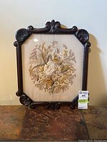 Front view of floral embroidery in a decorative dark carved wood frame displayed against a wall.