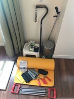 Photo showing orange yoga mat, two canes, small white plastic stool, heating pad, wrist weights, hand grips, resistance band expander, and metal trash bin on floor.