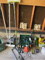Wide view of all items including broom, dustpan, hose, sprinklers, gloves, watering can, and house numbers in garage setting.