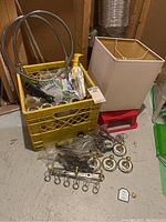 Photo showing yellow crate with various homeware items including flexible metal shower head hose, paint supplies, squares lamp shade, and metal hooks on floor