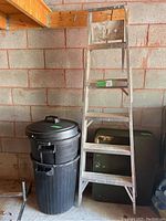 Photo showing 6ft Featherlite aluminum step ladder next to two black Rubbermaid trash bins with lids, against a concrete block wall in garage.