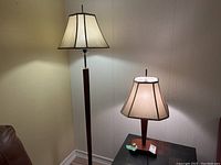 Photo showing full view of floor lamp and table lamp with beige fabric shades and wooden bases, lit to display color and style