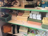 Shelf with neatly folded assorted towels including brown, beige, green, orange, and yellow. Plastic basket with folded linens and small sewing tools such as spools of thread.