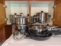 Full view of several stainless steel pots and pans of assorted sizes and some with lids, arranged inside a cabinet.
