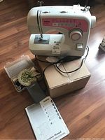 Photo showing the Brother XL-2610 sewing machine placed on its box with the box of sewing notions open beside it along with foot pedal and manual on wooden floor.