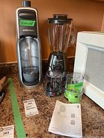 Full view of SodaStream soda maker with carbonation bottle, T-fal Blendforce blender with glass pitcher and attachments, and an instruction manual on a kitchen counter.