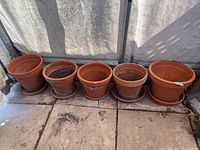 Five terracotta planters lined up showing different sizes and shapes, each paired with a terracotta saucer, placed on a stone floor against a fabric backdrop.