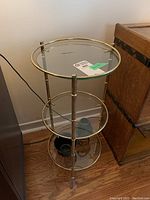 Full view of three tiered circular stand showing glass shelves and brass-colored metal frame with two decorative items on bottom shelf.