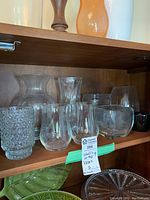 Eight clear glass vases displayed on a wooden shelf inside a cabinet, showing variety in size and texture, two matching vases visible