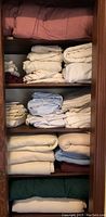Shelf with multiple folded sheets in various colors including white, cream, blue, burgundy, tan, and maroon, along with several folded blankets.