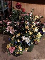 Wide angle photo showing a large group of assorted faux silk flowers in two green containers on the floor against a wood backdrop.