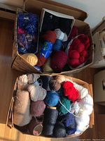 Two cardboard boxes filled with assorted yarn balls and skeins in different colors and textures, some loose and some boxed inside the bins.