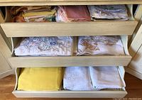 Three open drawers of a antique sideboard filled with folded embroidered and plain linen tablecloths and napkins in white, beige, yellow shades.