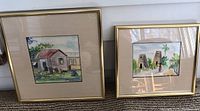 Two framed watercolor paintings by Vernon Grigg resting against white wall showing distinct Antigua scenes.
