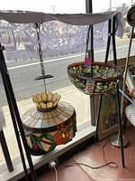 Two suspended stained glass ceiling light fixtures with floral and leaf patterns, hanging with chains. One has a wide bowl shape with wavy edges; the other has a conical form with a flower-shaped top piece.