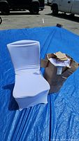 White stretch fabric chair cover fitted on a chair, showing front and side profile with a blue tarp and vehicles in the background.