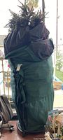 Full view of the green canvas carrying bag with the artificial tree tops protruding from the open bag, showing wheeled platform and black straps.
